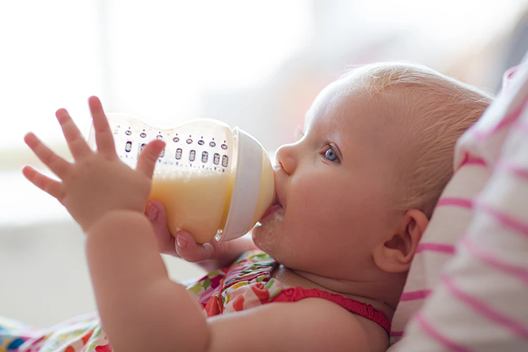 Blondes Baby mit blauen Augen und bunten Kleid trinkt ein Fläschen Milch
